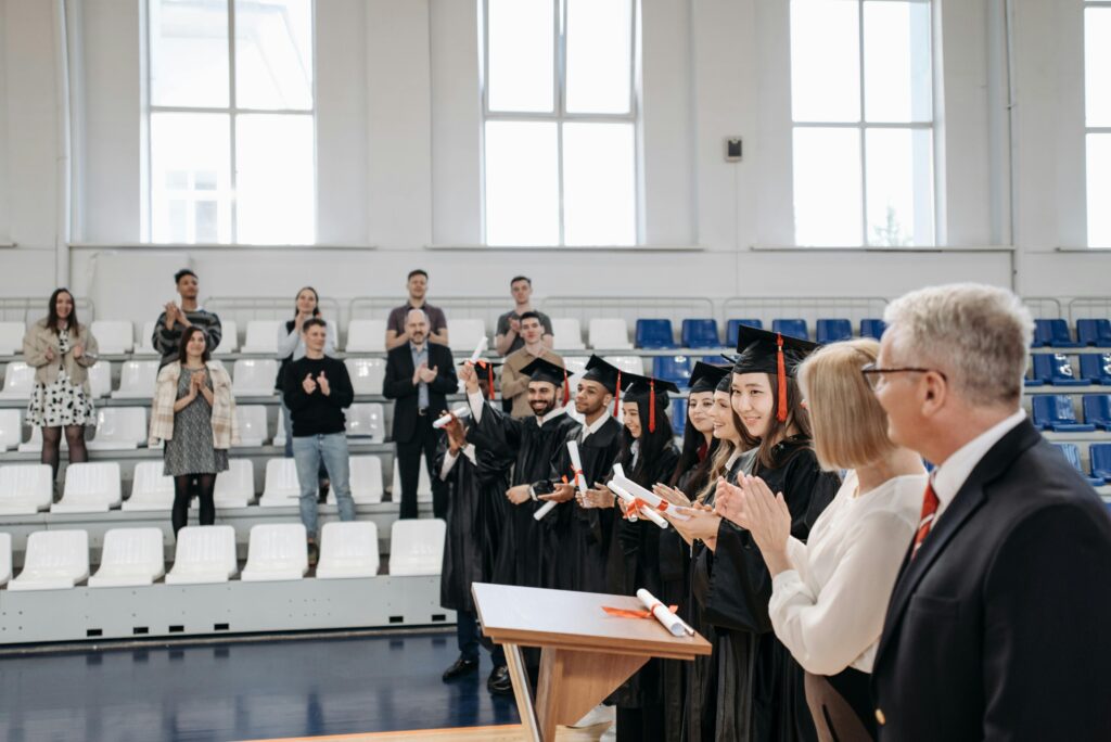 Graduates in gowns receive applause during an indoor ceremony, celebrating achievement.